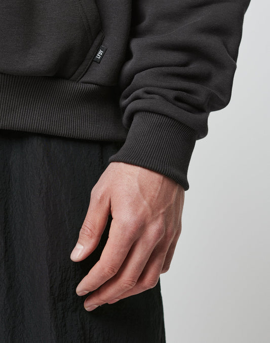 A close-up of a person’s left hand and wrist, wearing black textured pants and the oversized Hit and Run Hooded sweatshirt by LIVE FAST DIE YOUNG, featuring ribbed fabric and a small fabric tag near the waistband.