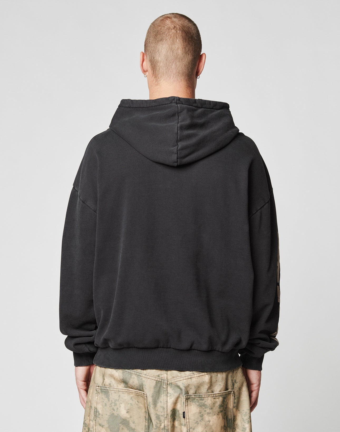 A person with a shaved head faces away from the camera, wearing a black LIVE FAST DIE YOUNG LFDY Angel Hooded and greenish-tan camo pants. The casual, relaxed look is set against a plain light gray background.