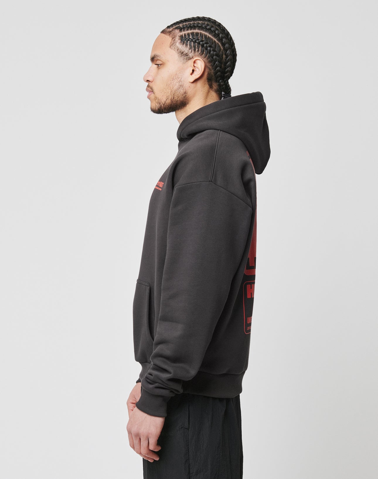 A man with braided hair, wearing the LIVE FAST DIE YOUNG Hit and Run Hooded oversized black hoodie with red graphics, stands in profile facing left against a plain white background.