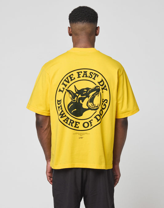 Someone is shown from behind wearing the LIVE FAST DIE YOUNG LFDY Circle Dog Tee, a bright yellow oversize fit t-shirt with a large circular graphic of a dog’s head and the text LIVE FAST, DIE YOUNG, BEWARE OF DOGS on the back. | model