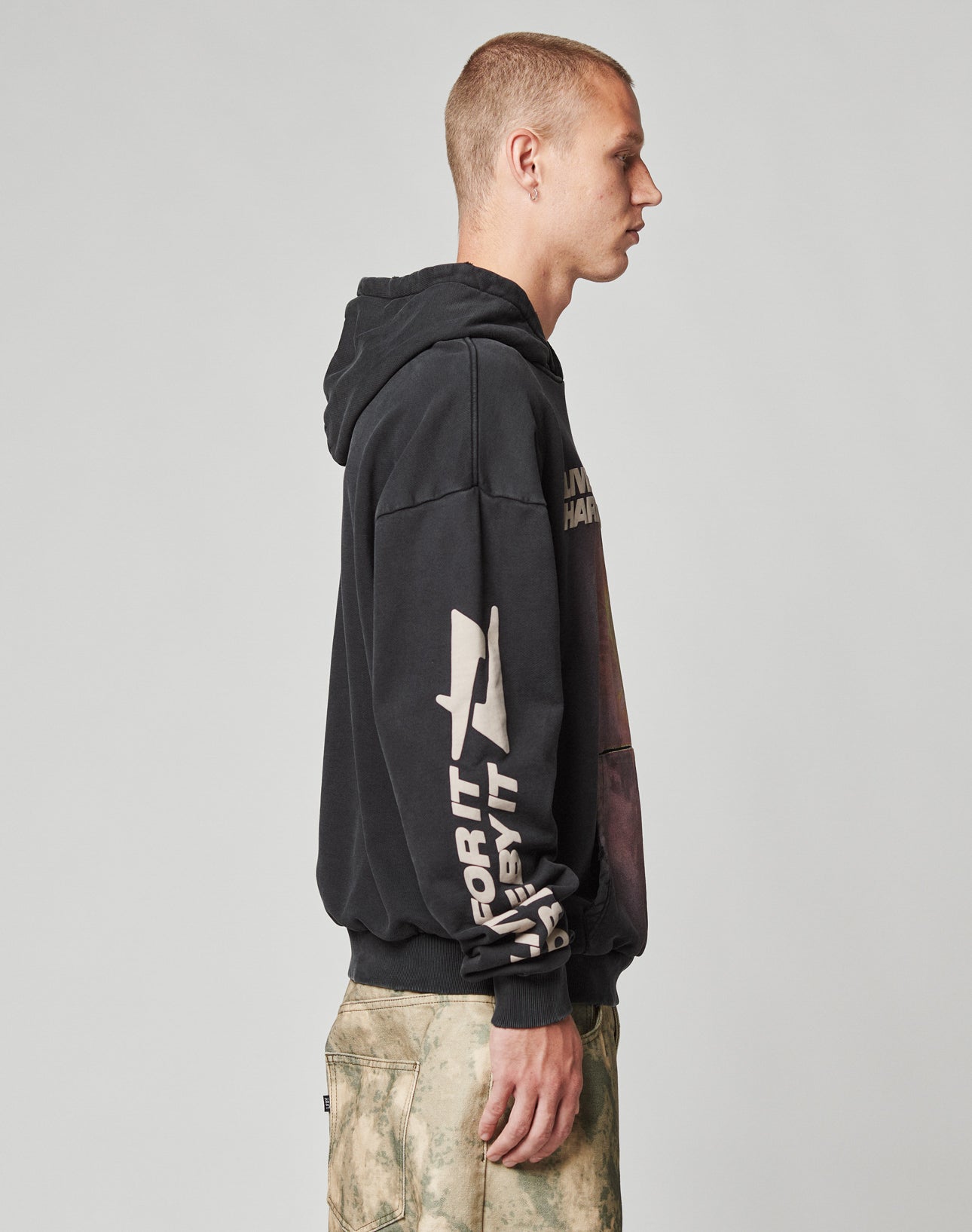 A young man with short hair stands in profile wearing a black LIVE FAST DIE YOUNG LFDY Angel Hooded hoodie with bold white and graphic text on the sleeve, and beige tie-dye pants, against a plain light gray background.