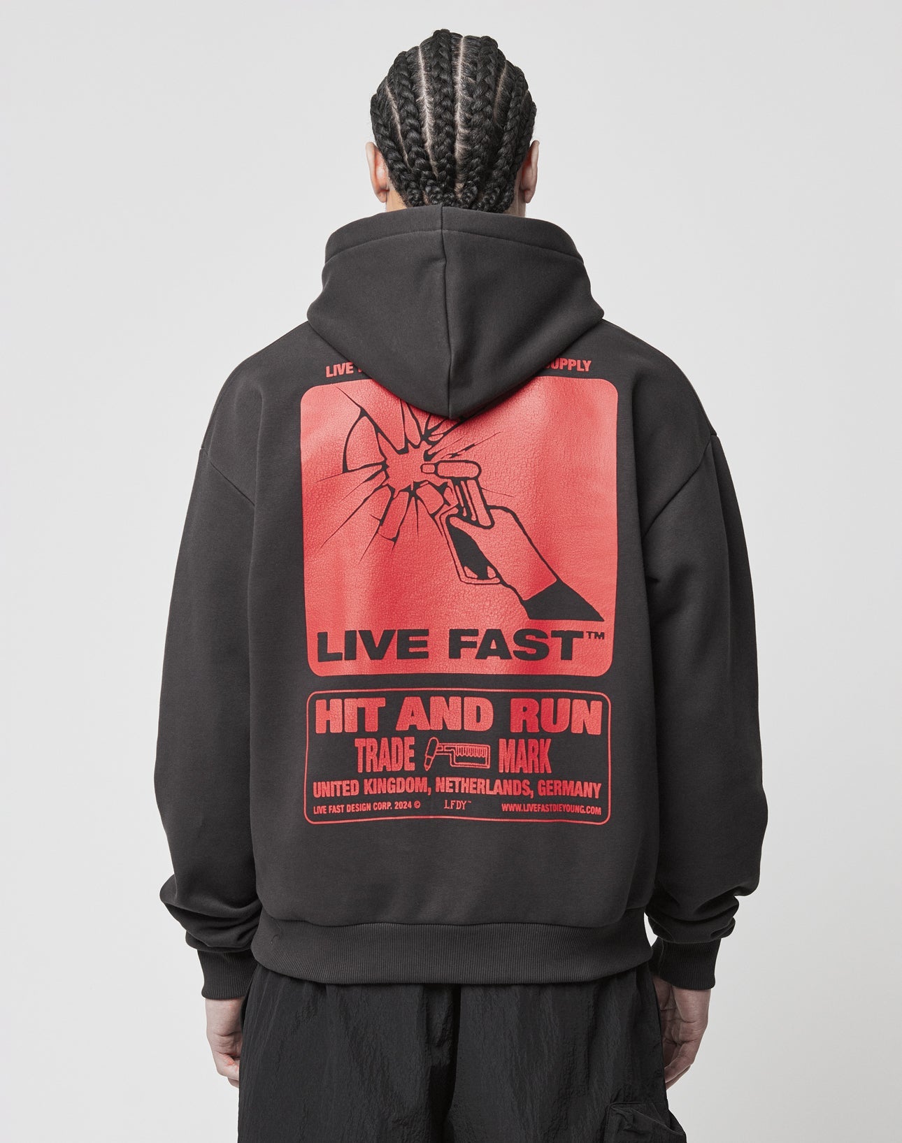 A person with braided hair wears the LIVE FAST DIE YOUNG Hit and Run Hooded—a black oversized hoodie with a large cracked backprint of a hand lighting a cigarette, bold “LIVE FAST” and “HIT AND RUN” text, plus smaller details.