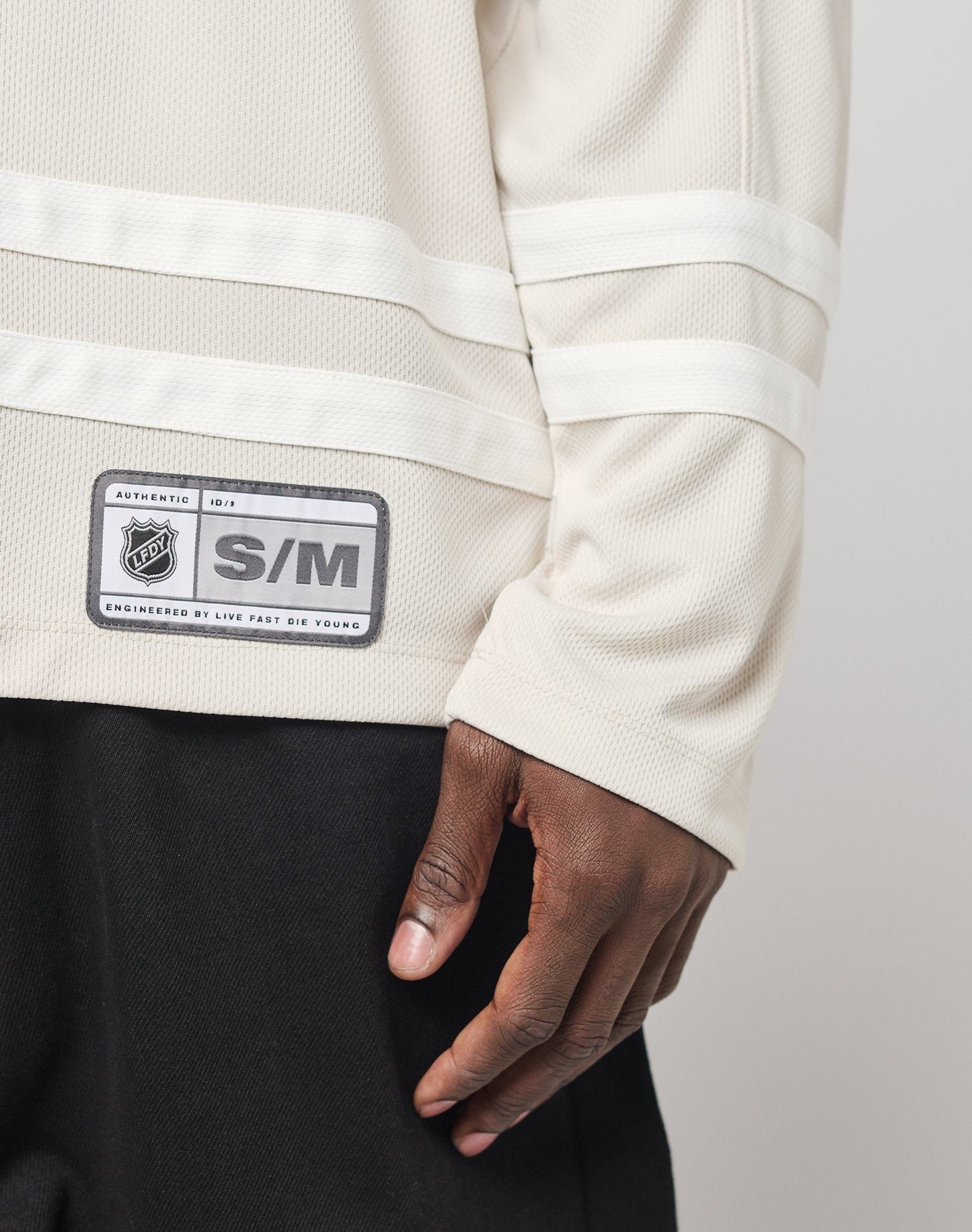 A person wears a cream LFDY Hockey Jersey by LIVE FAST DIE YOUNG in size S/M, featuring an oversized fit, over black pants. Only the lower part of the jersey and their hand at their side are visible.