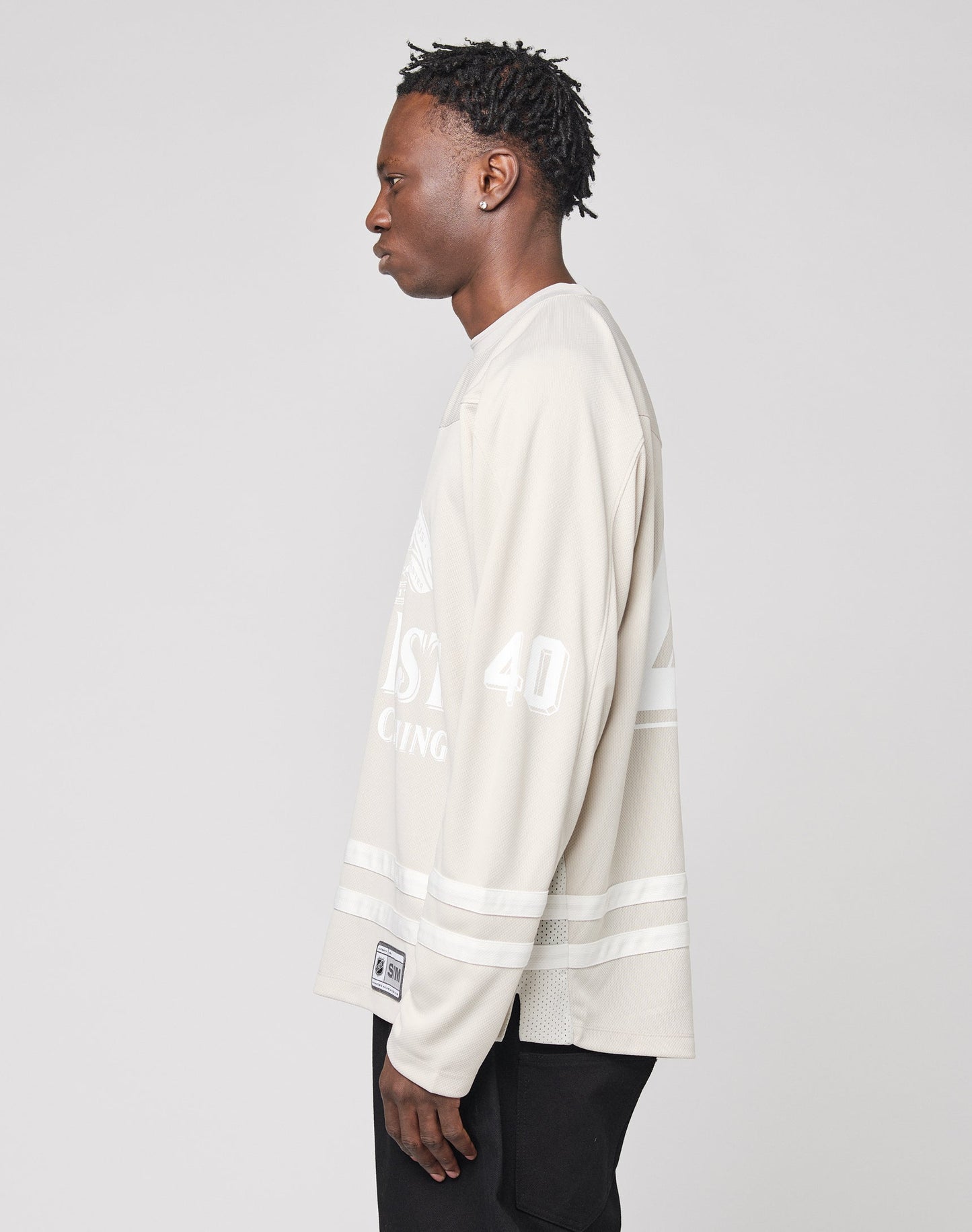 A person with short, twisted hair stands sideways, facing left, wearing the LIVE FAST DIE YOUNG LFDY Hockey Jersey—an oversize light beige shirt with a white sleeve print and number 40—paired with black pants against a plain background.