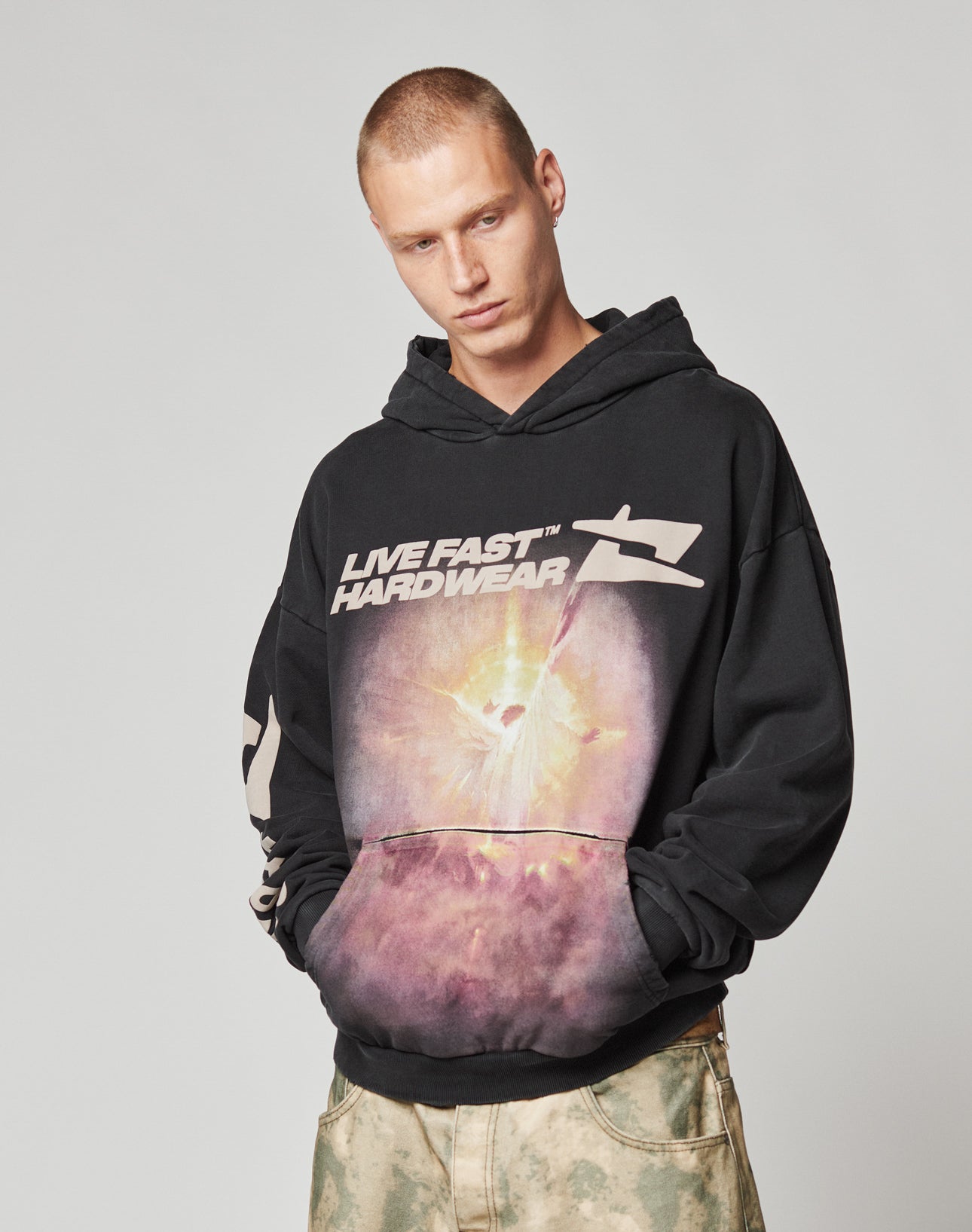 Someone in an oversize-fit LFDY Angel Hooded by LIVE FAST DIE YOUNG, featuring a cosmic graphic and LIVE FAST HARDWEAR text, pairs it with light tie-dye pants while standing against a plain background.
