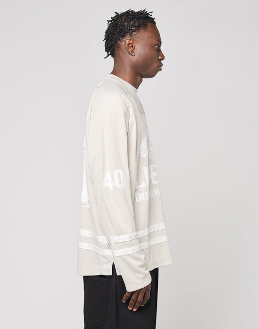 A person with short, textured hair stands in profile facing right, wearing the LIVE FAST DIE YOUNG LFDY Hockey Jersey—an oversize fit, light beige long-sleeve shirt with white graphics and Ärmelprint—paired with black pants.