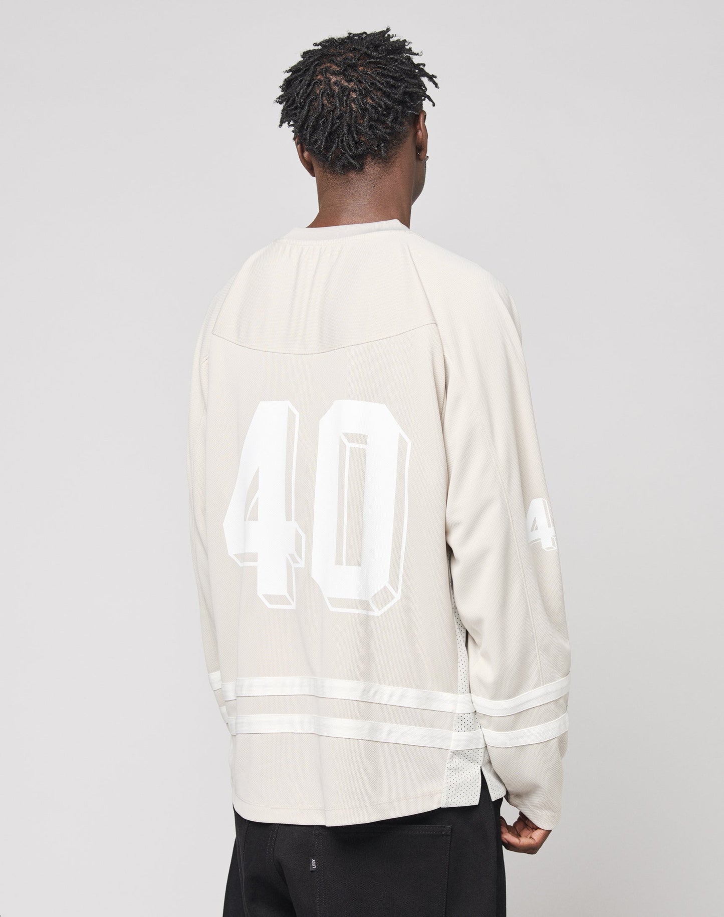 A person with short, twisted hair faces away, wearing the LIVE FAST DIE YOUNG LFDY Hockey Jersey in light beige, featuring an oversize fit and large white 40 print on the back and sleeve, paired with black pants.