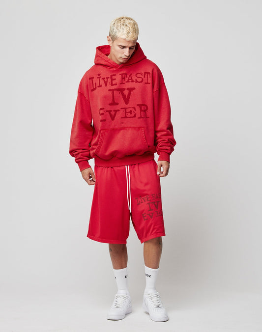 A young man with short blond hair wears LIVE FAST DIE YOUNG’s red Live Fast IV Ever Shorts with an elastic waistband and side pockets, styled with a matching hoodie, white socks, and sneakers against a gray background.
