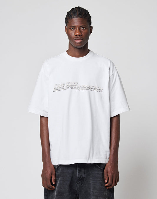 A young man with braided hair faces forward, wearing the LIVE FAST DIE YOUNG LFDY Europe Run Tee—a loose, oversized white t-shirt with embossed text—paired with dark jeans against a plain light gray background.