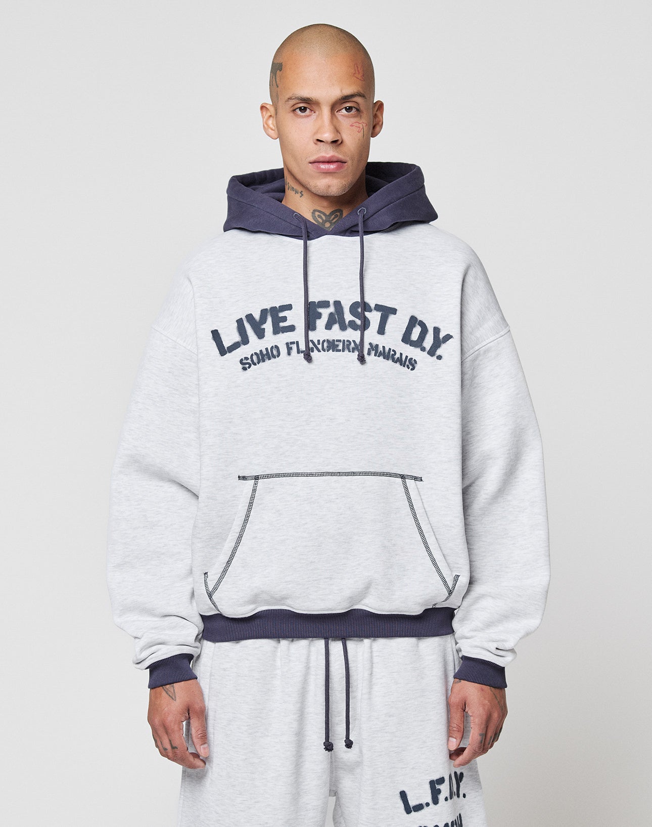 A person with a shaved head wears the LFDY District Hooded by LIVE FAST DIE YOUNG, with dark blue contrast drawstrings, a kangaroo pocket, and logo print. They pair it with matching gray sweatpants. | model