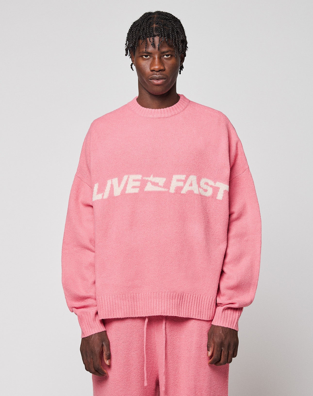 A person with short, twisted hair wears the LIVE FAST DIE YOUNG Live Fast Boucle Sweater in an oversize pink fit with “LIVE FAST” in bold white letters, paired with matching pants against a plain, light background.