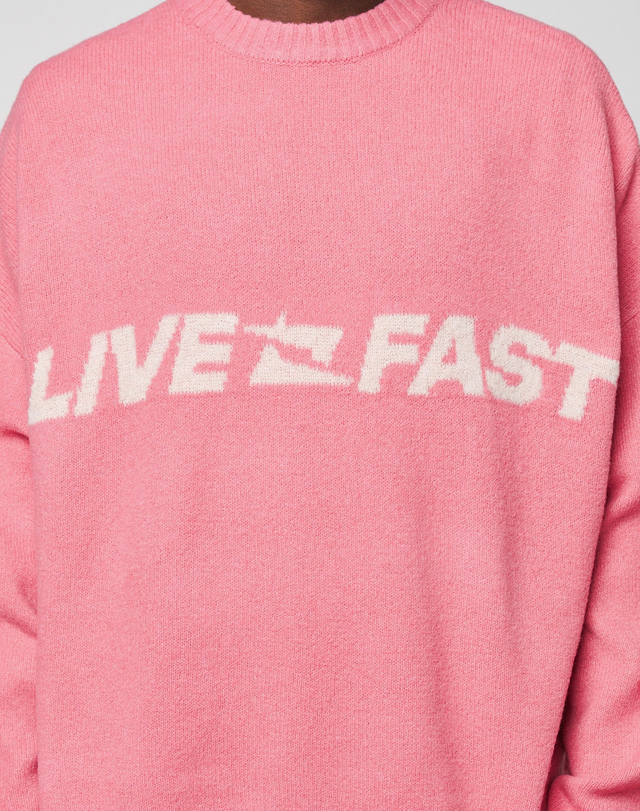 Someone wears the Live Fast Boucle Sweater by LIVE FAST DIE YOUNG—an oversize pink knit featuring "LIVE FAST" in bold white lettering across the chest.