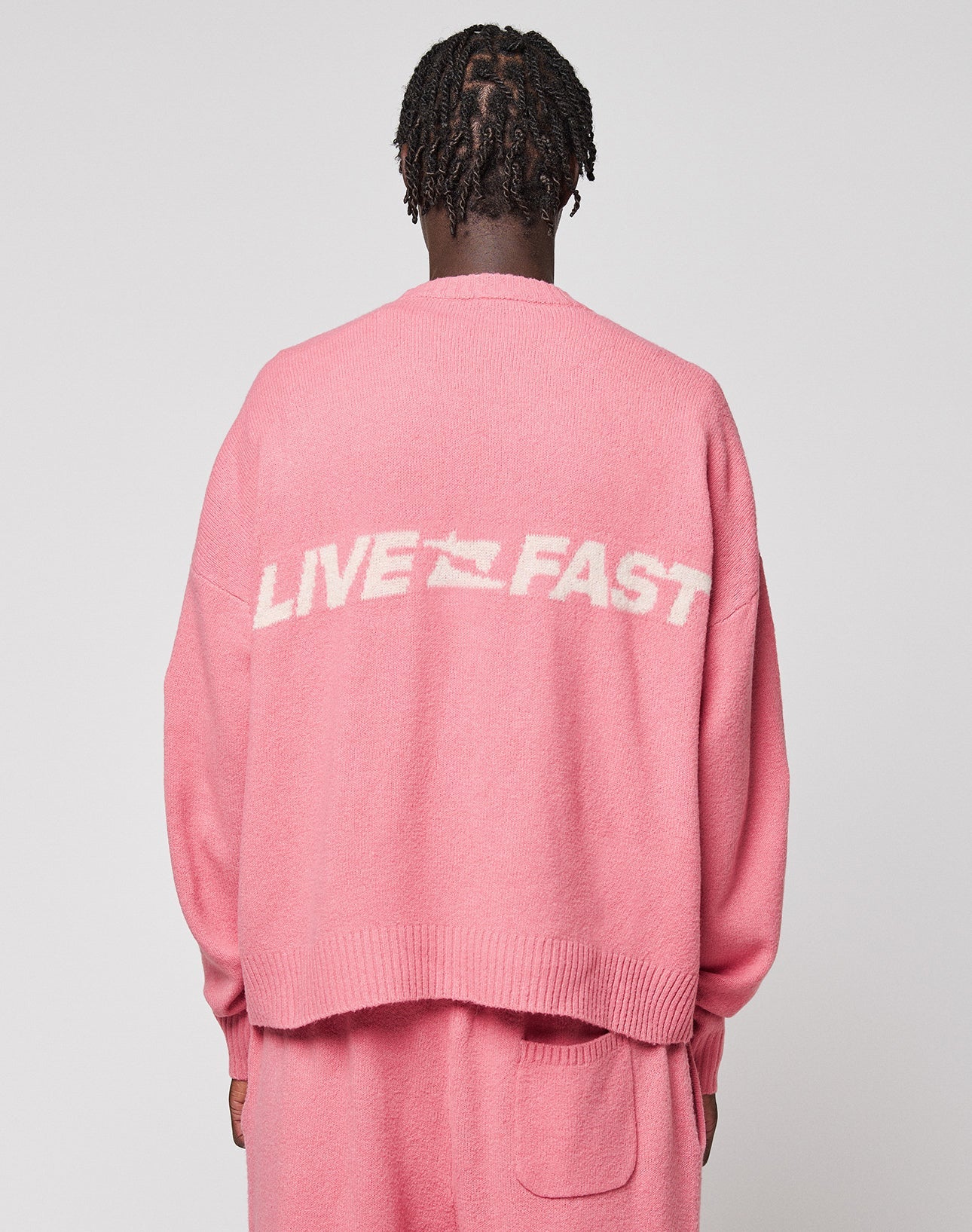 Seen from behind, a person with short braided hair wears the LIVE FAST DIE YOUNG "Live Fast Boucle Sweater," an oversized pink knit with "LIVE FAST" in white on the back, paired with matching pink pants against a plain light background.
