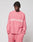 A person wears the LIVE FAST DIE YOUNG Live Fast Boucle Sweater in oversize-fit pink with “LIVE FAST” in white, paired with matching pink pants, standing hands in pockets and looking down against a plain light background.