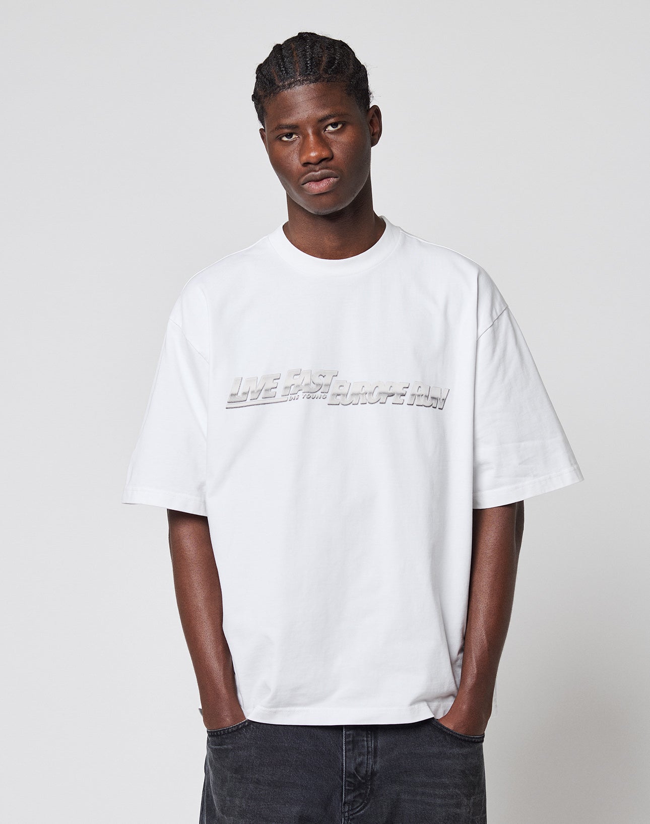 A young man with braided hair wears the LIVE FAST DIE YOUNG LFDY Europe Run Tee, an oversized white T-shirt with LIVE FAST embossed in large text, paired with dark pants, standing against a plain light gray background.
