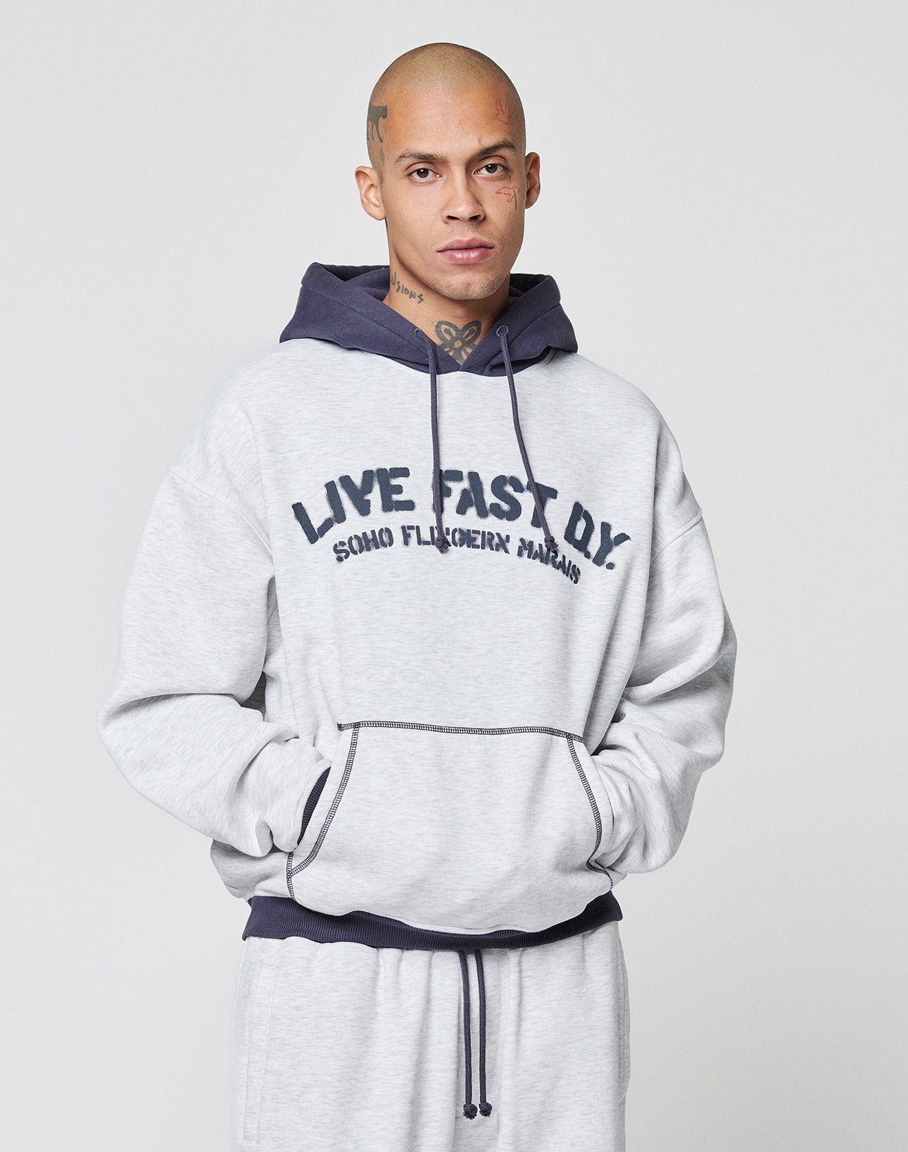 A person with a shaved head wears the LFDY District Hooded by LIVE FAST DIE YOUNG in light gray with contrast trim and the words LIVE FAST D.Y. SOHO FLUCREHX MAHAL printed on the front, against a plain light background.