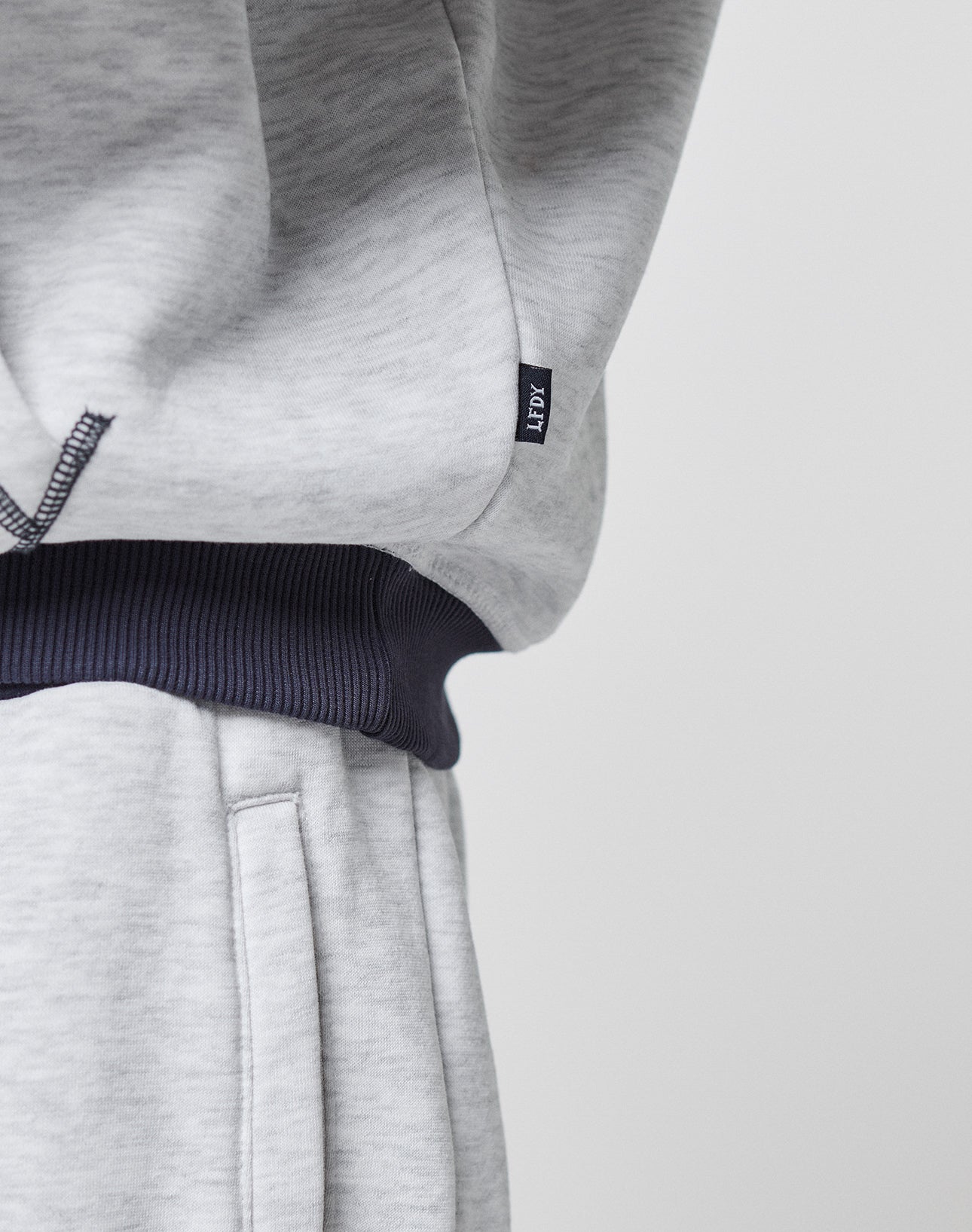 Close-up of a person in light gray LFDY District Hooded sweatpants and matching oversize hoodie by LIVE FAST DIE YOUNG, featuring a contrasting ribbed waistband and a small black tag with white lettering on the side seam.