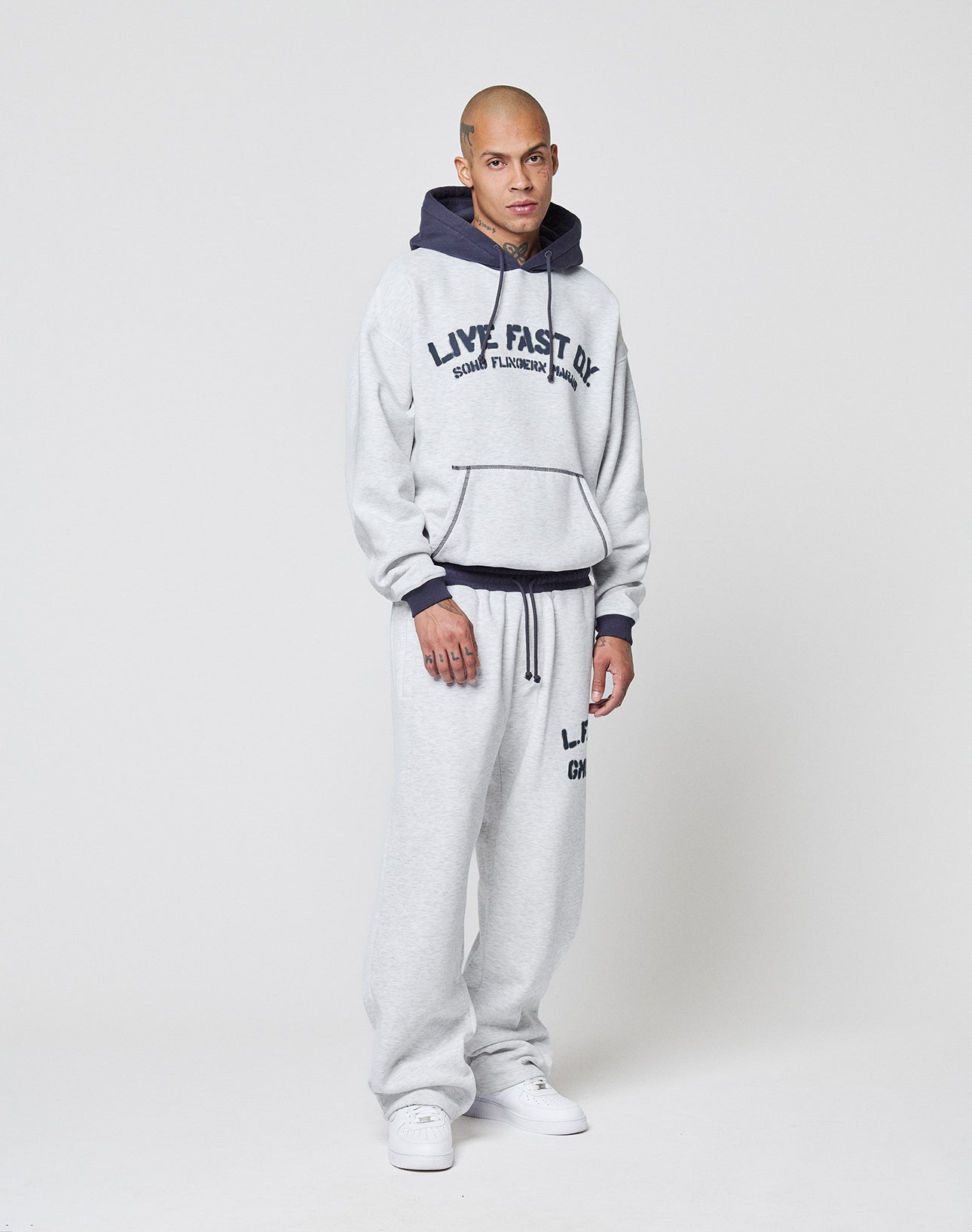 A man stands against a plain white background wearing LIVE FAST DIE YOUNG's LFDY District Jogger and matching light gray wide fit hoodie with "LIVE FAST" text, dark drawstrings, zippered side pockets, and white sneakers.