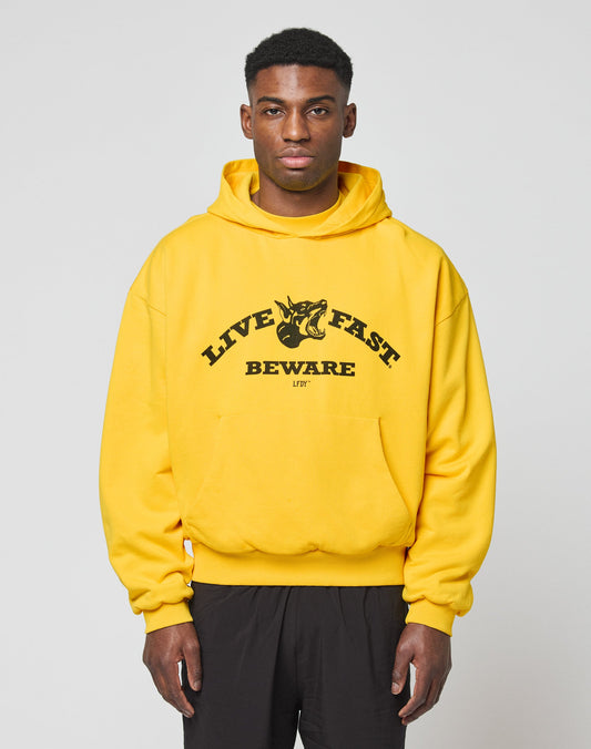A man wearing a bright yellow LIVE FAST DIE YOUNG Live Fast Beware Hooded with LIVE FAST BEWARE and a fierce animal graphic on the front stands against a plain light gray background.
