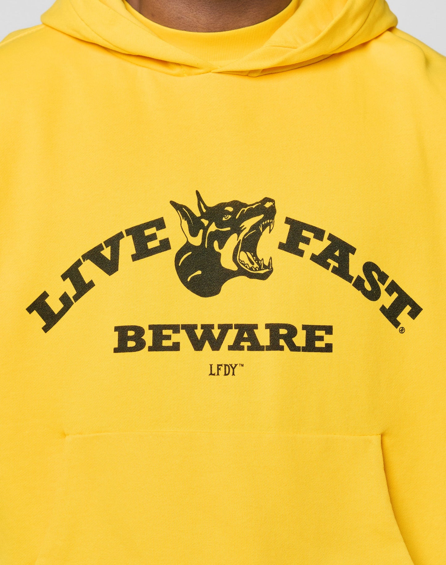 The LIVE FAST DIE YOUNG Live Fast Beware Hooded is a plain yellow oversize fit hoodie, laid flat facing backward on a light gray background. It features long sleeves, double layer 100% Baumwolle fabric, and no visible logos or designs.