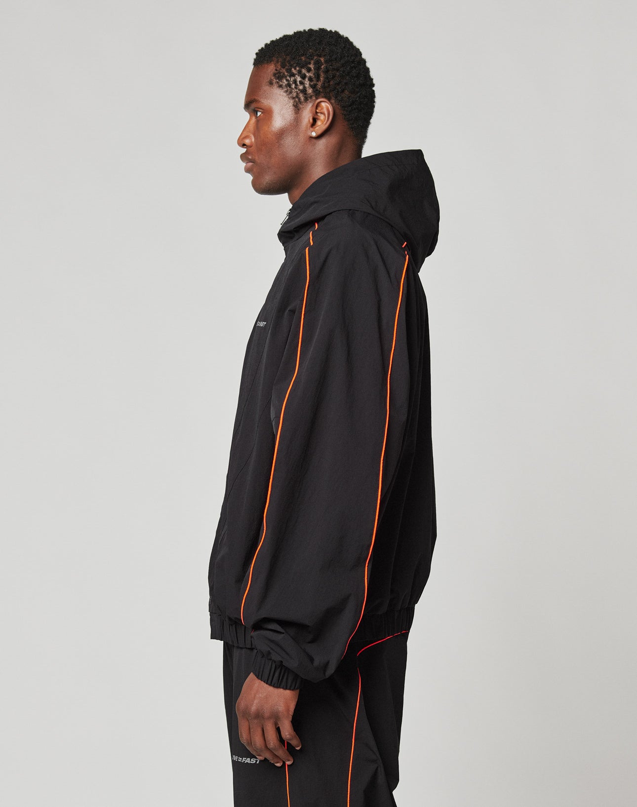 A man stands in profile wearing the LIVE FAST DIE YOUNG Live Fast Trackjacket, a black hooded nylon jacket with orange piping and elastic cuffs, paired with matching black pants against a plain light background.