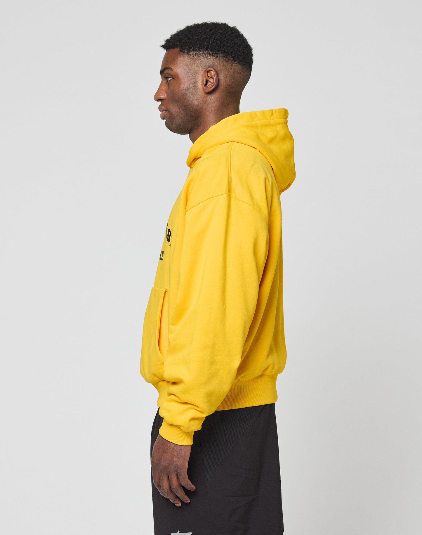 A man stands in profile facing left, wearing a bright yellow LIVE FAST DIE YOUNG Live Fast Beware Hooded and black shorts against a plain light gray background.