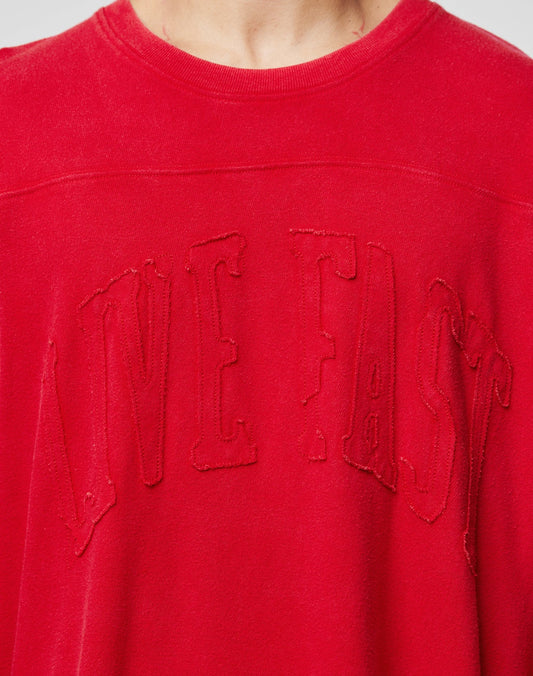 A close-up of a person wearing a red Ripped College Longsleeve by LIVE FAST DIE YOUNG, featuring the brand’s name embroidered in large, slightly frayed letters across the chest.