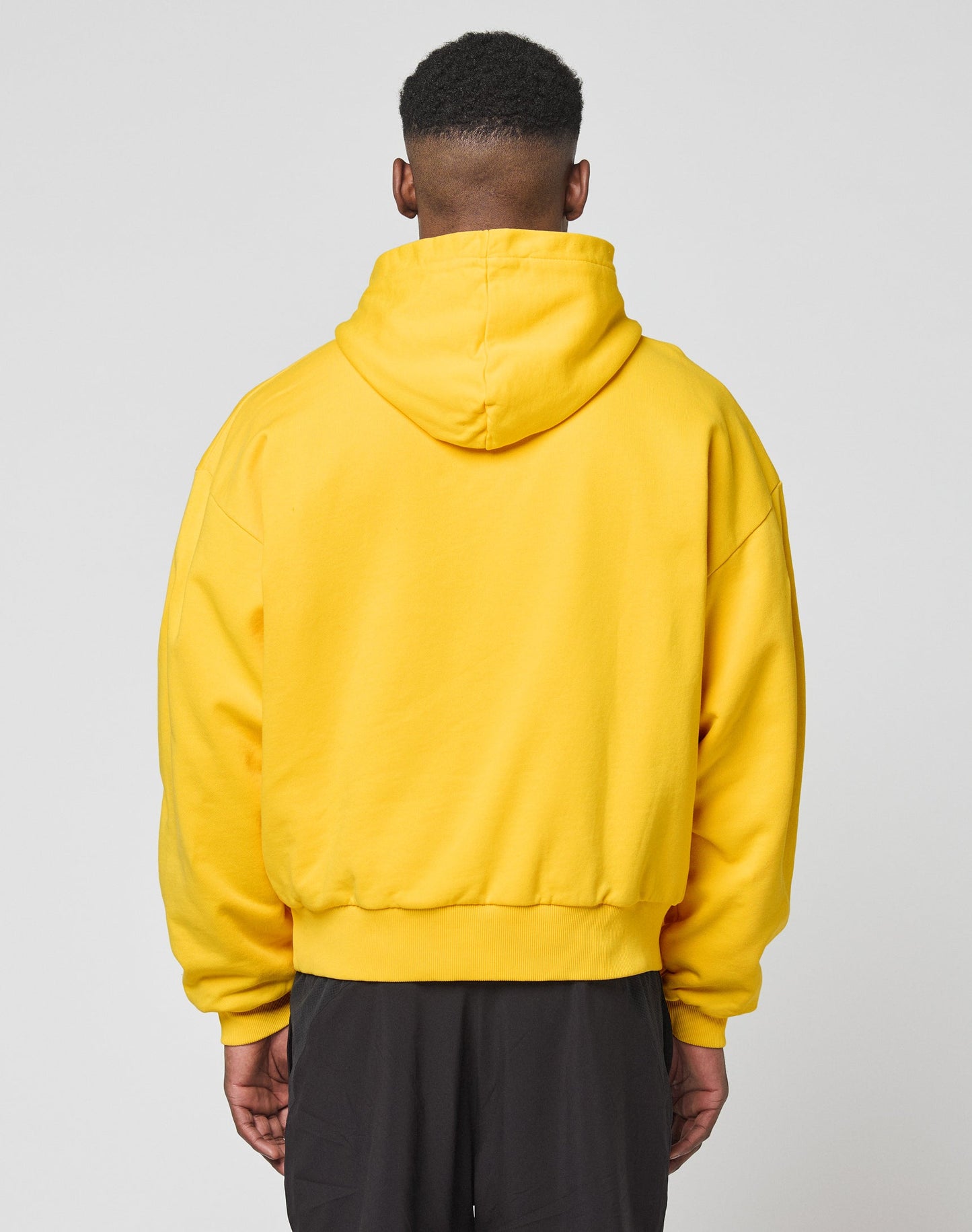A person faces away from the camera, wearing a bright yellow LIVE FAST DIE YOUNG Live Fast Beware Hooded and dark pants against a plain light gray background.