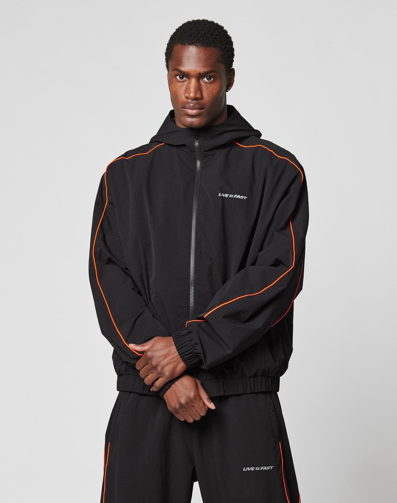 A man wears the LIVE FAST DIE YOUNG Live Fast Trackjacket—black nylon with orange piping, reflective LIVE FAST print, and elastic cuffs—standing against a gray background, looking serious with hands clasped.