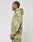 A man with cornrows stands in profile wearing the LIVE FAST DIE YOUNG Camo Hooded Zip, featuring a green and beige splatter pattern, along with matching sweatpants, against a plain light background.