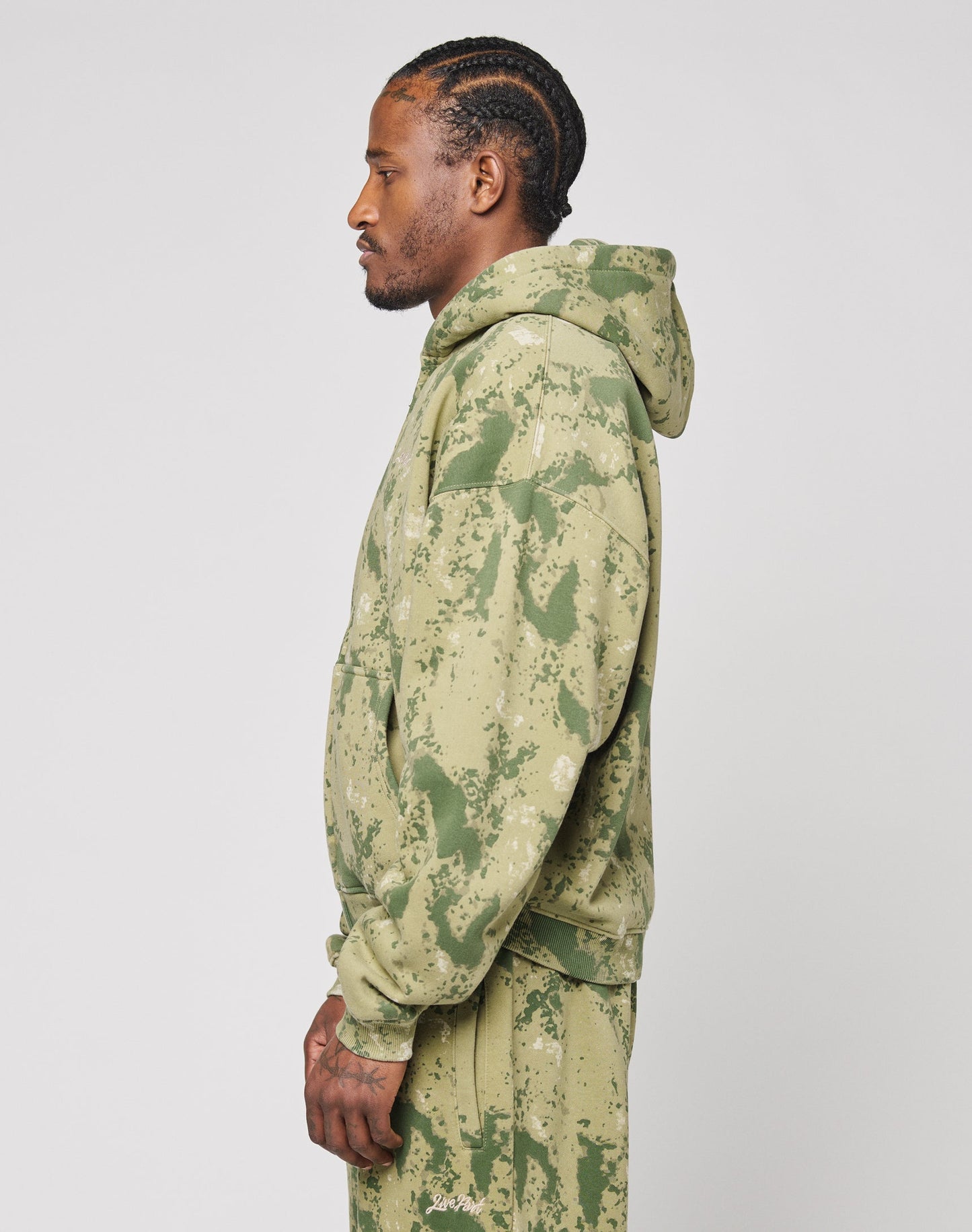 A man with cornrows stands in profile wearing the LIVE FAST DIE YOUNG Camo Hooded Zip, featuring a green and beige splatter pattern, along with matching sweatpants, against a plain light background.