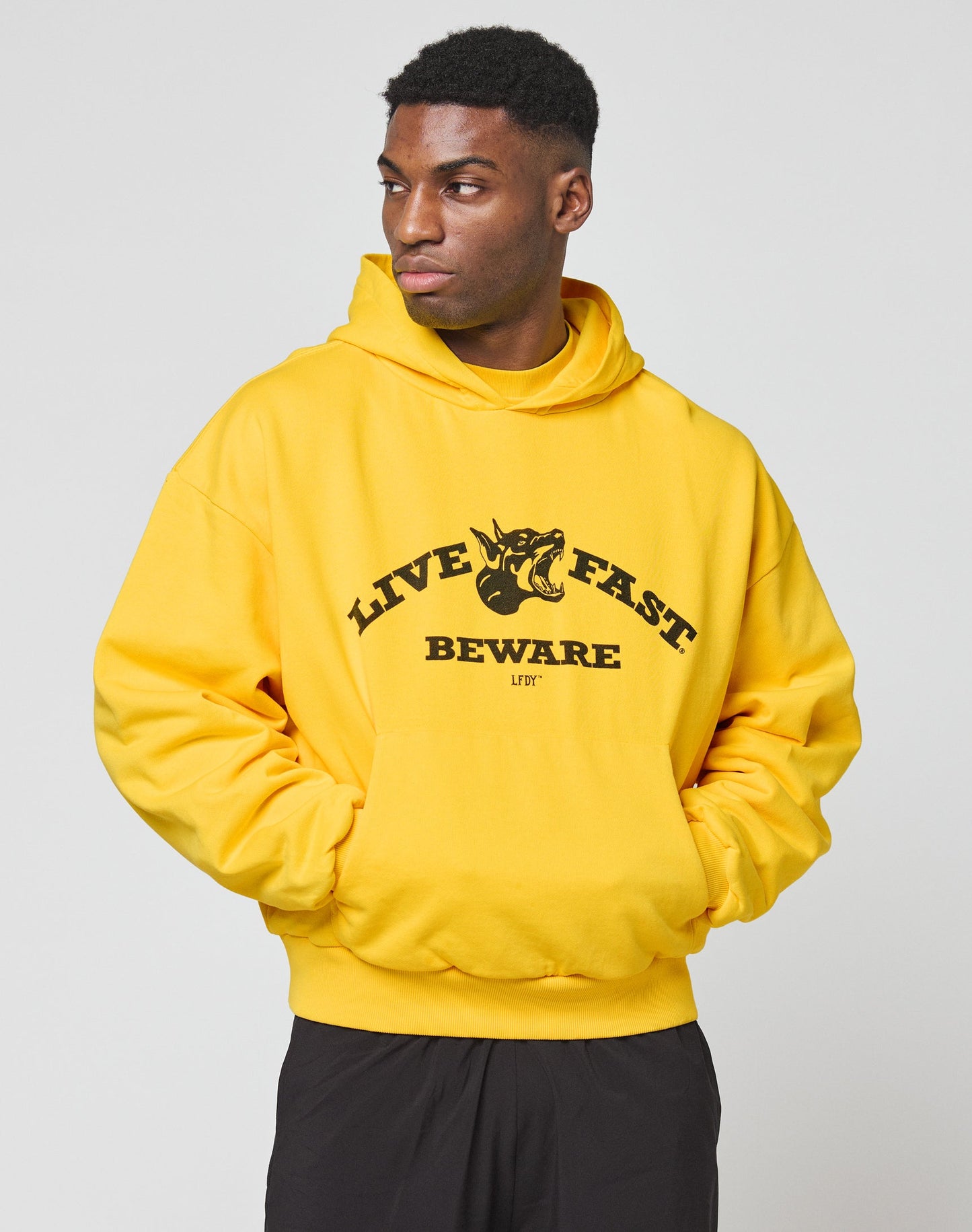 Someone in a bright yellow LIVE FAST DIE YOUNG Live Fast Beware Hooded, featuring a dogs head graphic and the text LIVE FAST BEWARE on the front, stands with hands in pockets, gazing to the side.