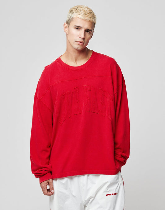 A person with short blond hair wears the LIVE FAST DIE YOUNG Ripped College Longsleeve in red, featuring distressed appliqué, paired with matching white shorts, standing against a plain light gray background.