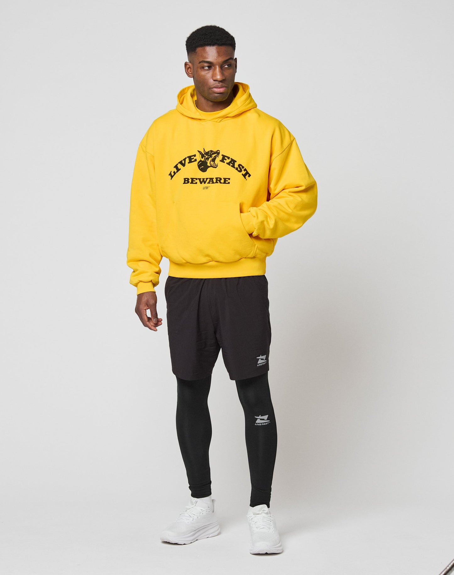 A man stands against a plain background wearing the LIVE FAST DIE YOUNG Live Fast Beware Hooded in bright yellow, paired with black shorts, black leggings, and white sneakers.