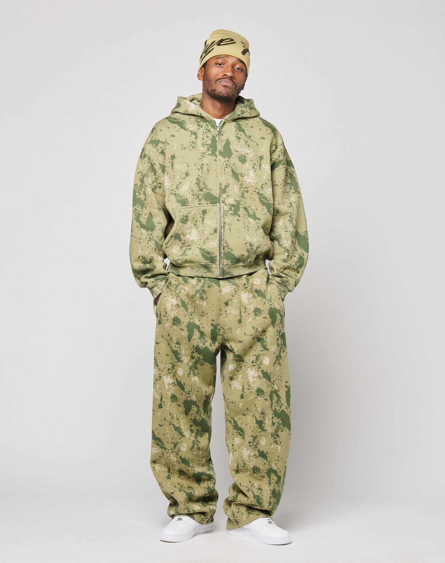 A man stands against a plain background wearing the LIVE FAST DIE YOUNG Camo Hooded Zip in Oversize Fit with matching pants, a yellow Nike headscarf, and white sneakers, hands in pockets and a neutral expression.