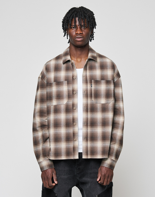 A man with medium-length, twisted hair wears the LIVE FAST DIE YOUNG Vintage Flannel—an oversized brown and beige plaid shirt layered over a white tee—while standing against a plain gray background. | model