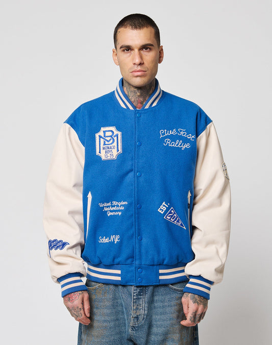 A man with short hair and tattoos wears the LIVE FAST DIE YOUNG LFDY x Schott NYC Varsity Jacket with embroidered patches, cowhide leather sleeves, and Monaco Rallye text over light blue jeans, standing against a plain white background. | model