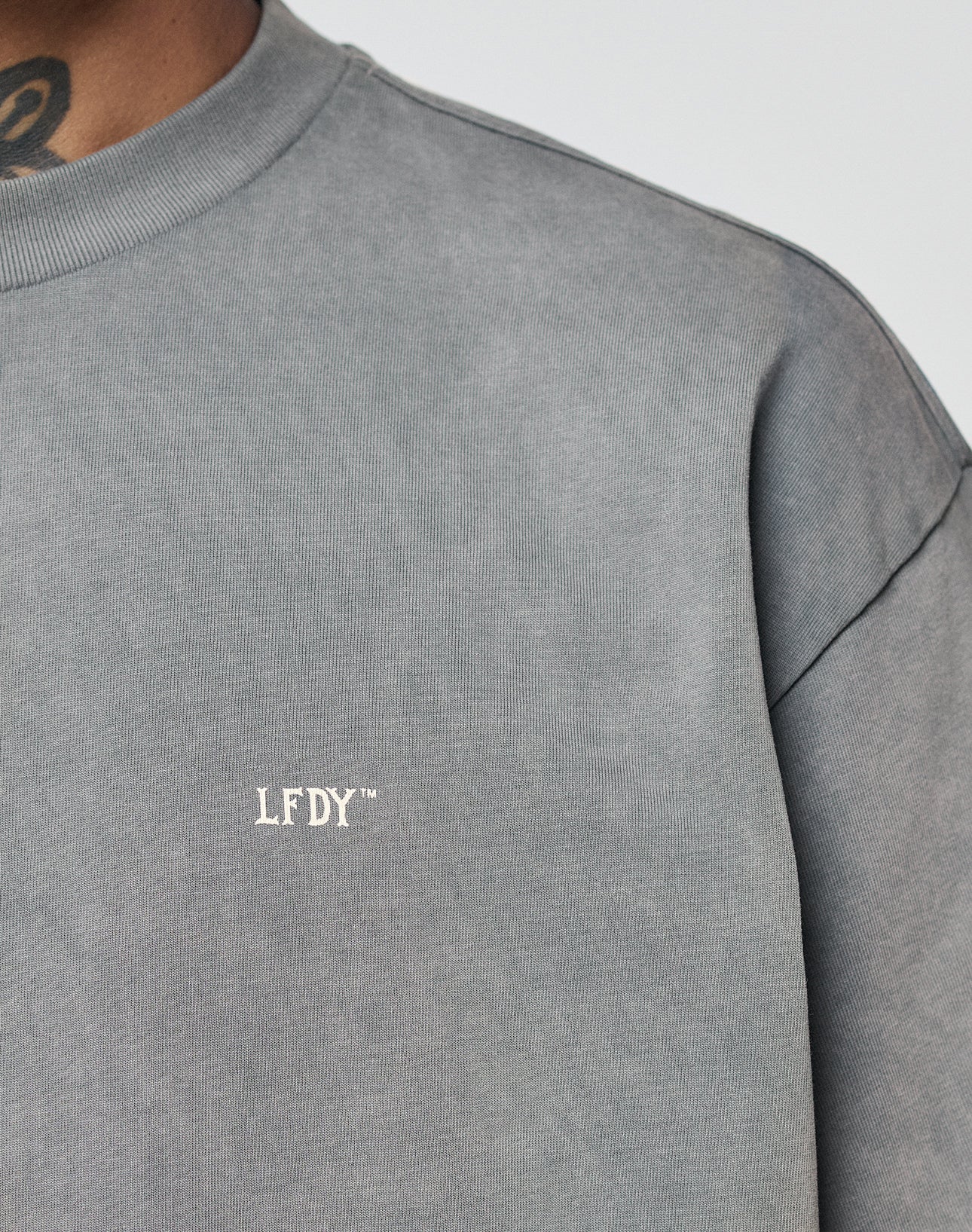 Close-up of a person wearing the grey Basic Trademark Tee by LIVE FAST DIE YOUNG, with small white LFDY™ embroidery on the chest. Part of the persons neck and a tattoo are partially visible.
