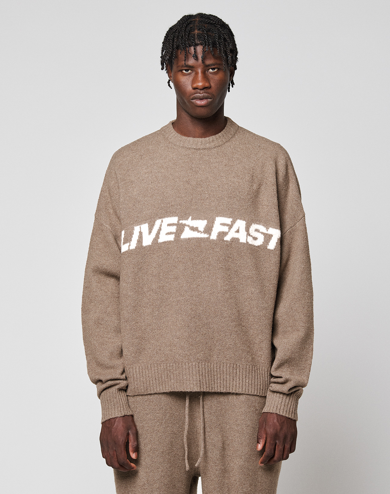 A person wears the LIVE FAST DIE YOUNG Live Fast Boucle Sweater, an oversized beige knit with "LIVE FAST" in bold white letters, paired with matching pants against a plain background. | model