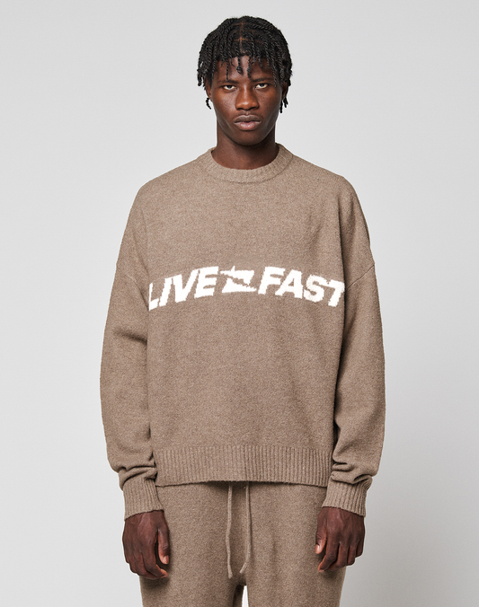 A person wears the LIVE FAST DIE YOUNG Live Fast Boucle Sweater, an oversized beige knit with "LIVE FAST" in bold white letters, paired with matching pants against a plain background. | model