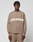 A person wears the LIVE FAST DIE YOUNG Live Fast Boucle Sweater, an oversized beige knit with "LIVE FAST" in bold white letters, paired with matching pants against a plain background.