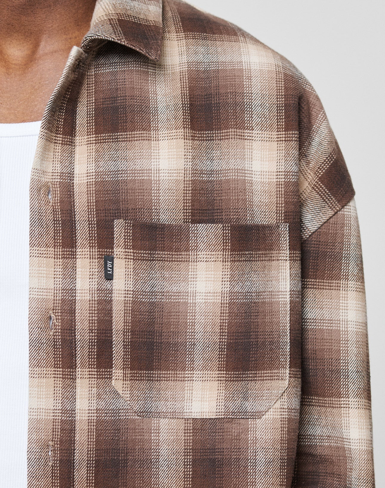 A close-up shows a person wearing the LIVE FAST DIE YOUNG Vintage Flannel in brown and beige plaid over a white ribbed undershirt. Only the chest and neck are visible, with an LFDY-branded front pocket on a small black tag.