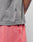 A person wearing a gray Basic Trademark Tee by LIVE FAST DIE YOUNG with a small black tag and pink shorts stands with one hand in their pocket, revealing part of their colorful arm tattoo.