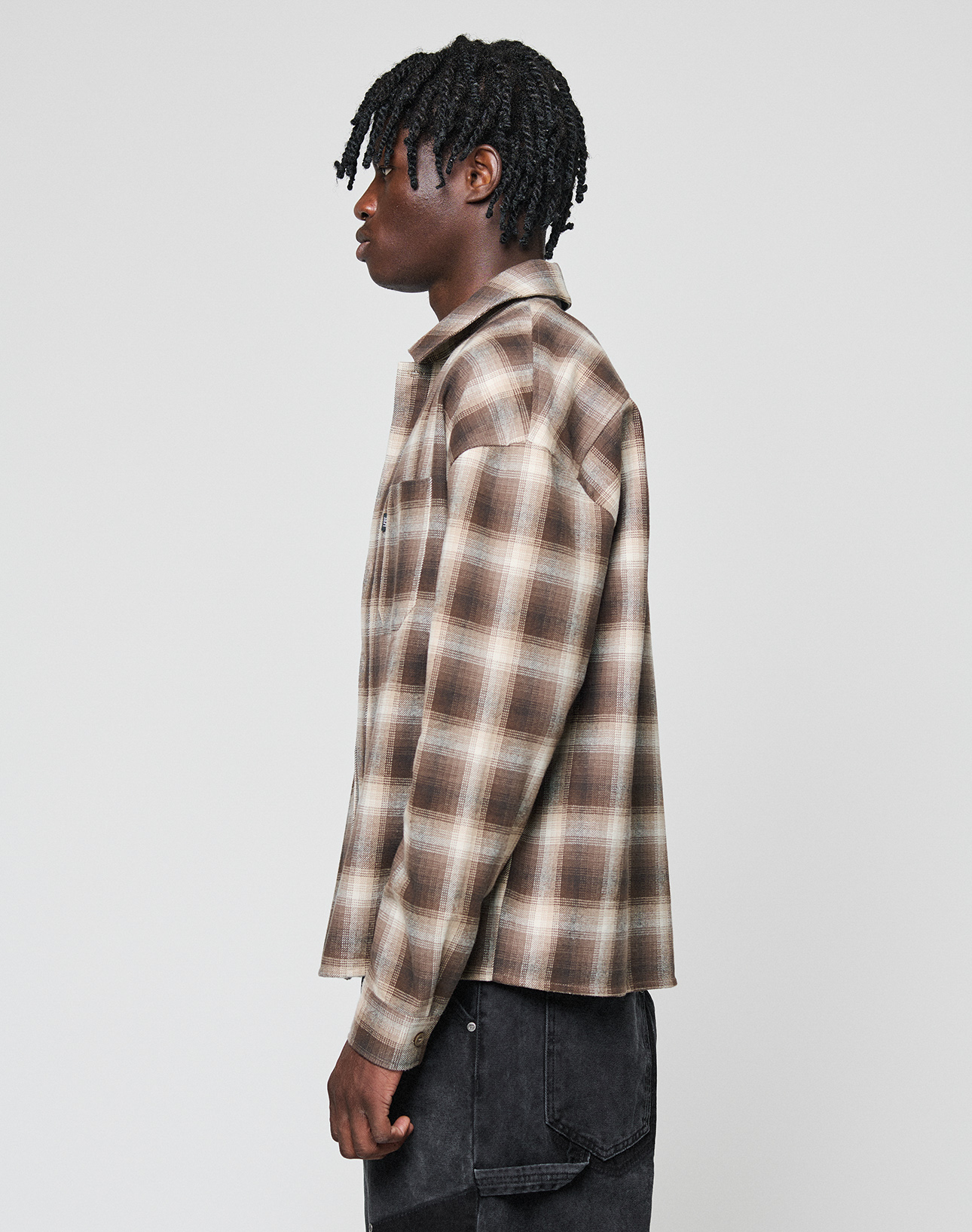 A person with short, twisted hair in profile sports a LIVE FAST DIE YOUNG Vintage Flannel—an oversized brown and beige plaid shirt made of 100% Baumwolle—paired with black jeans, set against a plain light gray backdrop.
