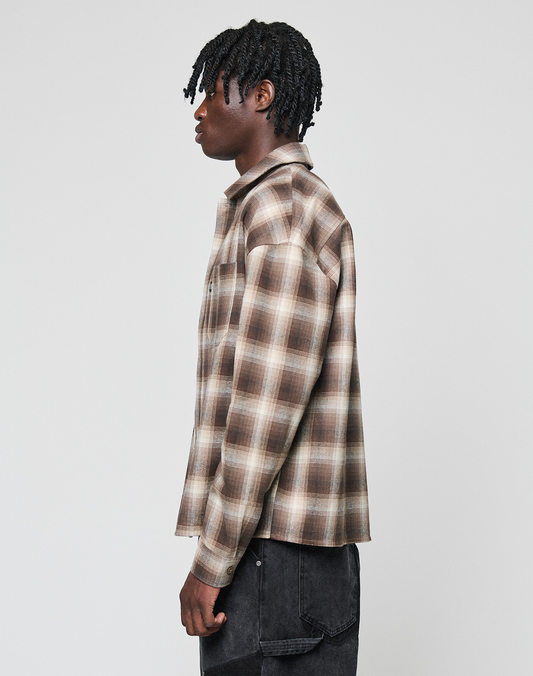 A person with short, twisted hair in profile sports a LIVE FAST DIE YOUNG Vintage Flannel—an oversized brown and beige plaid shirt made of 100% Baumwolle—paired with black jeans, set against a plain light gray backdrop.
