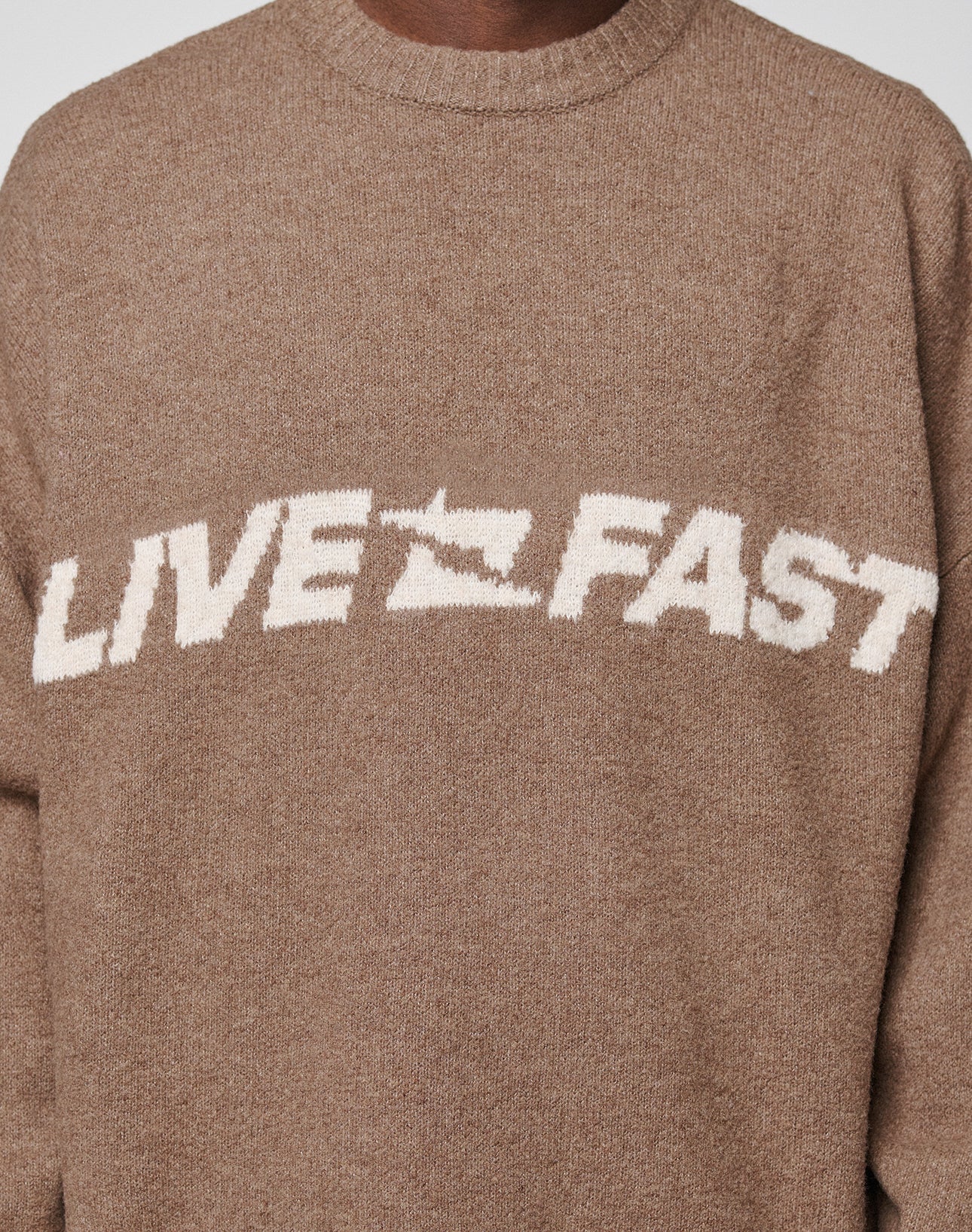 A person wears the Live Fast Boucle Sweater by LIVE FAST DIE YOUNG in brown, featuring "LIVE FAST" in bold white letters across the chest, with the 'A' in FAST uniquely stylized as a star.