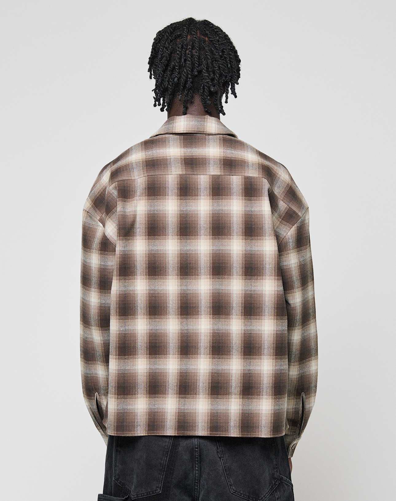 A person with short twists is shown from behind wearing the LIVE FAST DIE YOUNG Vintage Flannel in brown and beige plaid with black pants, oversize fit, against a plain white background.