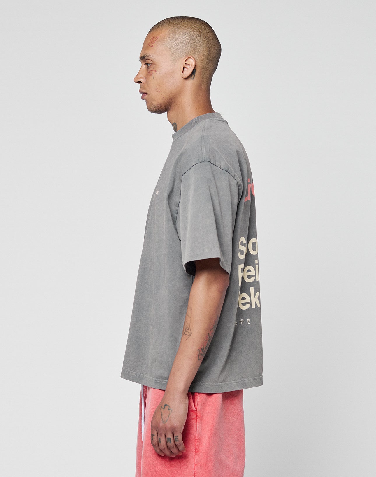 A man with a shaved head stands sideways, wearing the oversize gray Basic Trademark Tee aus Baumwolle with front and back print by LIVE FAST DIE YOUNG and pink shorts. The background is plain and light-colored.