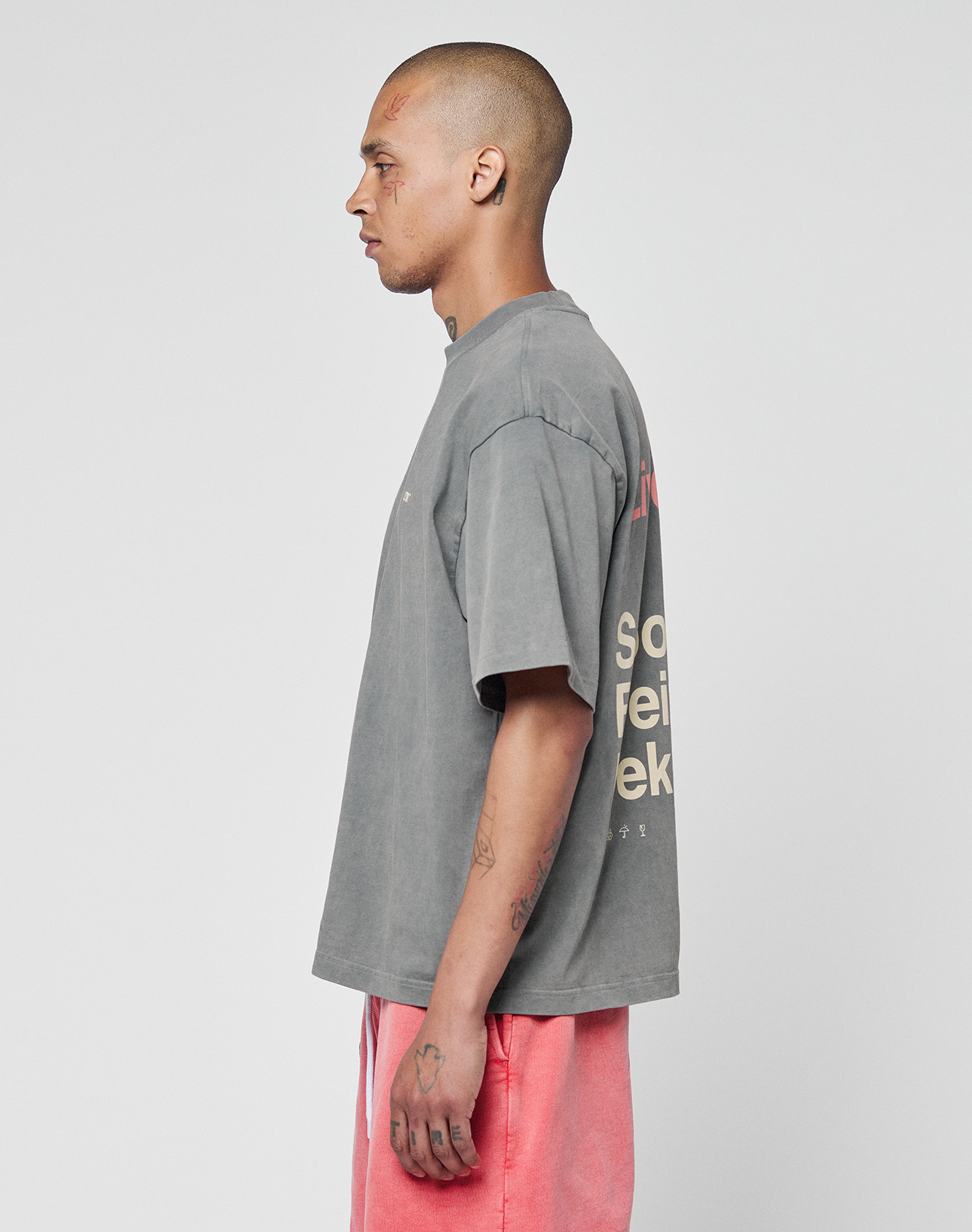 A person with a shaved head and facial tattoos stands in profile, wearing the Basic Trademark Tee by LIVE FAST DIE YOUNG in gray oversize fit with back text and pink shorts, their unique look contrasting the plain white background.