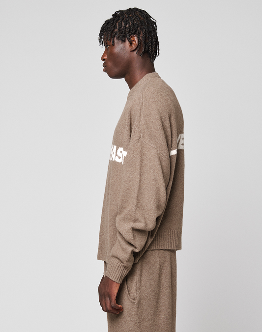 A person with short, textured hair stands sideways against a plain light gray background, wearing the Live Fast Boucle Sweater by LIVE FAST DIE YOUNG in an oversize fit with matching beige pants.